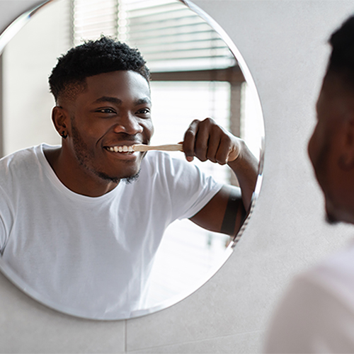 African Guy Cleaning Teeth Smiling To In Mirror In Bathroom